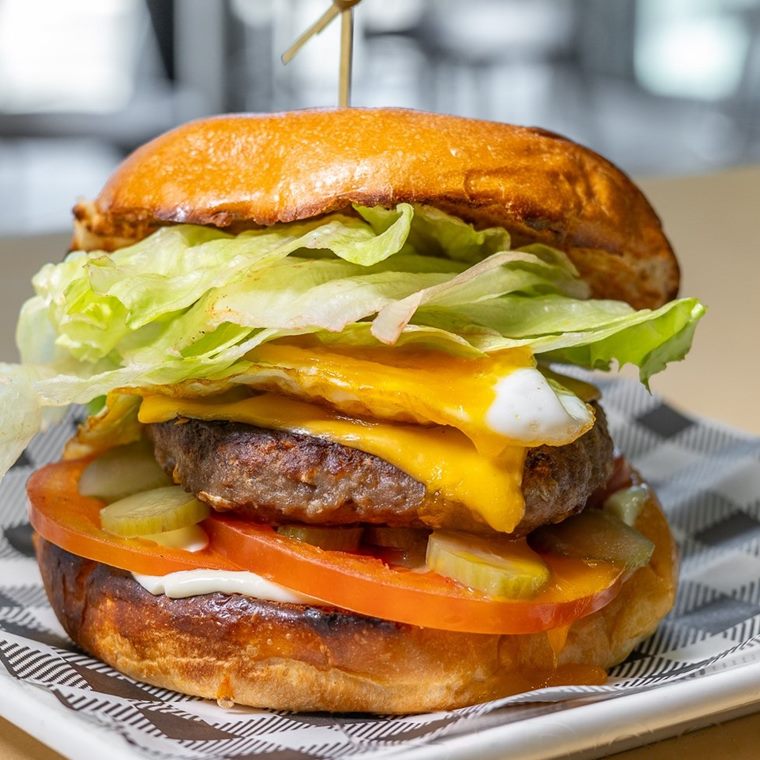 A hamburger with beef patty, egg, American cheese, lettuce, pickles and tomato in a brioche bun.